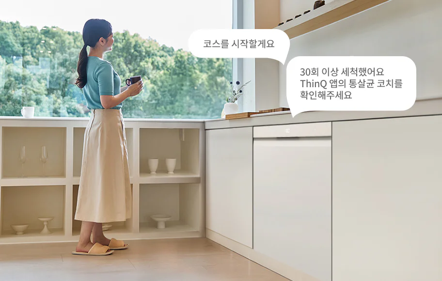 주방에서 한 여성이 창밖을 바라보며 머그잔을 들고 서 있습니다. 식기세척기 위쪽에는 코스를 시작할게요라는 텍스트가 표시되어 있으며, 그 옆에는 30회 이상 세척했어요. ThinQ 앱의 통살균 코치를 확인해주세요라는 문구가 있습니다. 창가에는 화병과 컵이 놓여 있으며, 창 밖으로는 푸른 나무들이 보입니다.